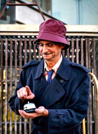 El Flaco, de El Flaco Circus, con sombrero burdeos y gabardina oscura, tocando un timbre de recepción con gesto concentrado.