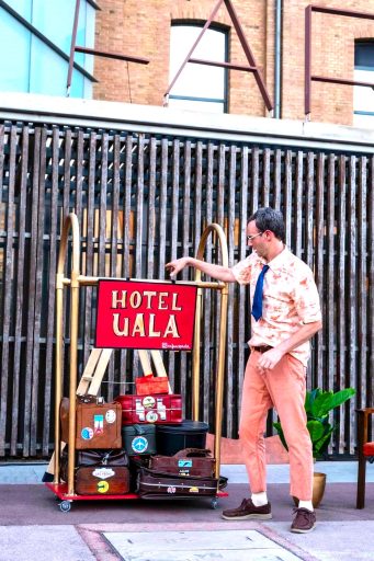 El Flaco, de El Flaco Circus, interactuando con el cartel del Hotel Ualá, junto a una estructura con maletas decoradas con pegatinas de destinos.