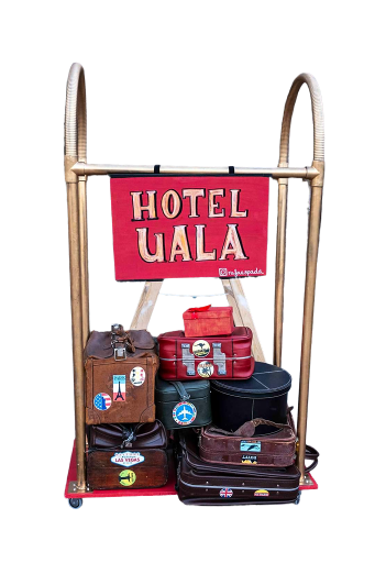 Estructura móvil con cartel rojo de “Hotel Ualá” y varias maletas decoradas con pegatinas de ciudades y destinos turísticos.